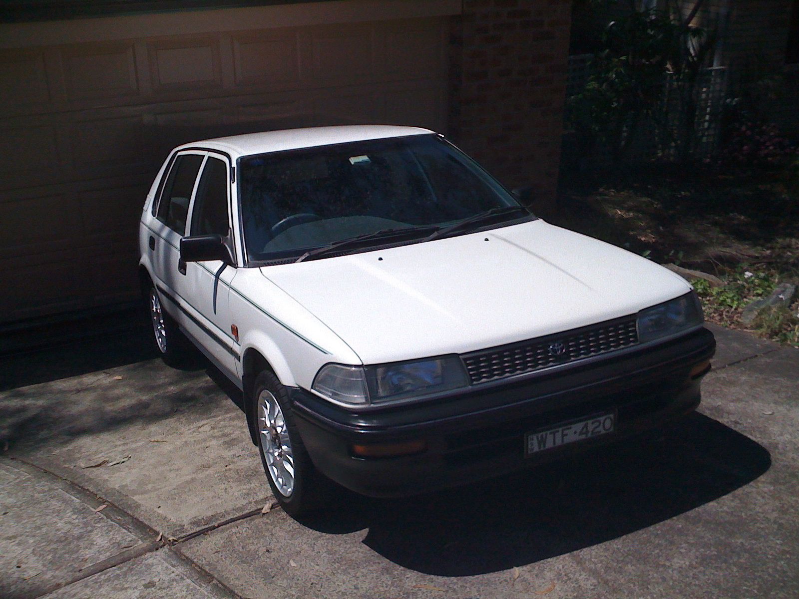 A photo of a white 1993 Toyota Corolla hatchback with low-profile tyres, lace-style rims, and numberplates that say "WTF420". It's very shiny and clean-looking.