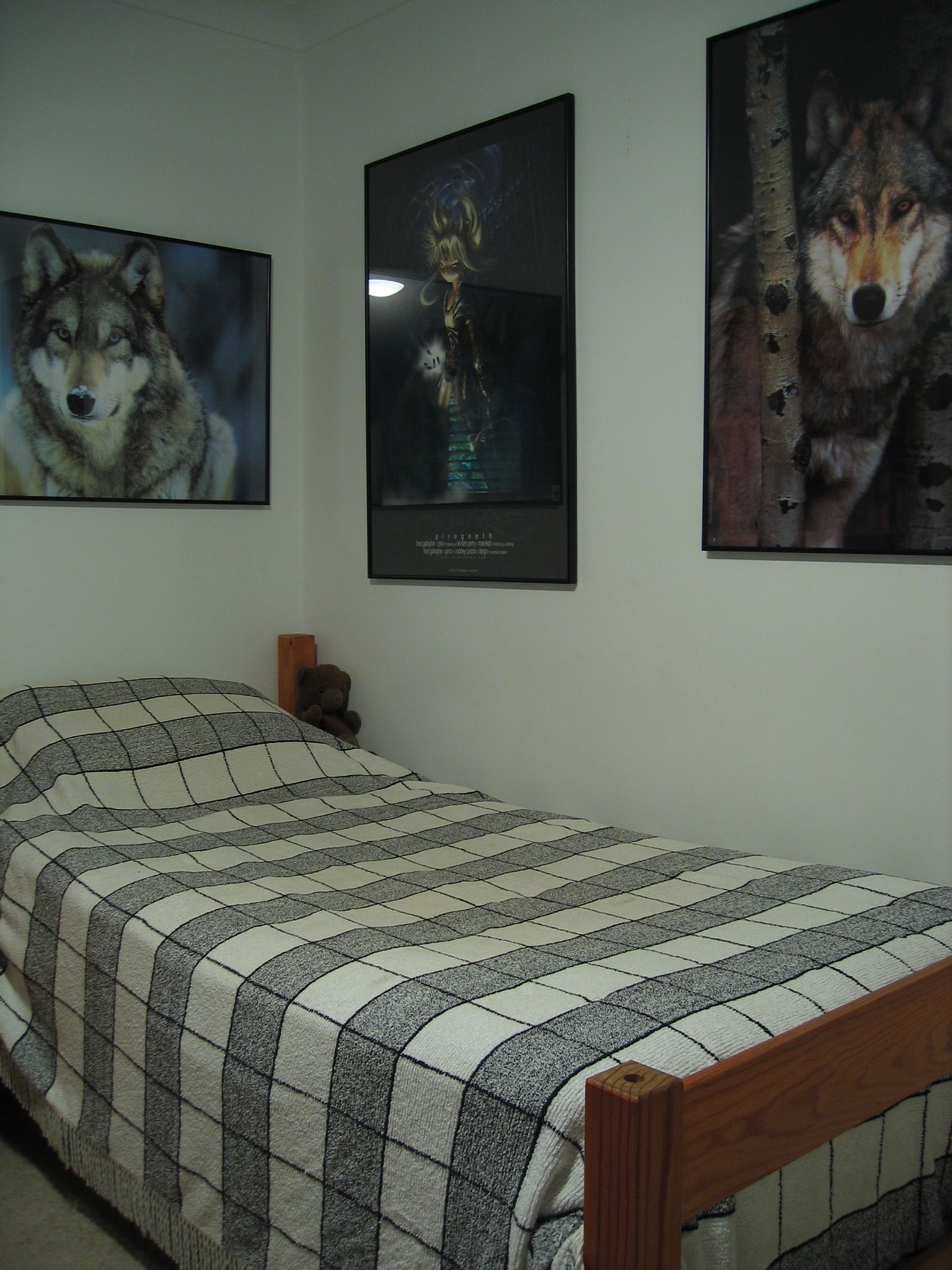 A photo of a single bed with a black and white square-pattern blanket on it, with three posters on the wall next to it. One is the aforementioned wolf poster, another is an ominous anime-style girl with blonde hair hovering just above the ground with a ball of light glowing in one hand, and the third is another wolf poster, this one a grey wolf standing next to a birch tree trunk.