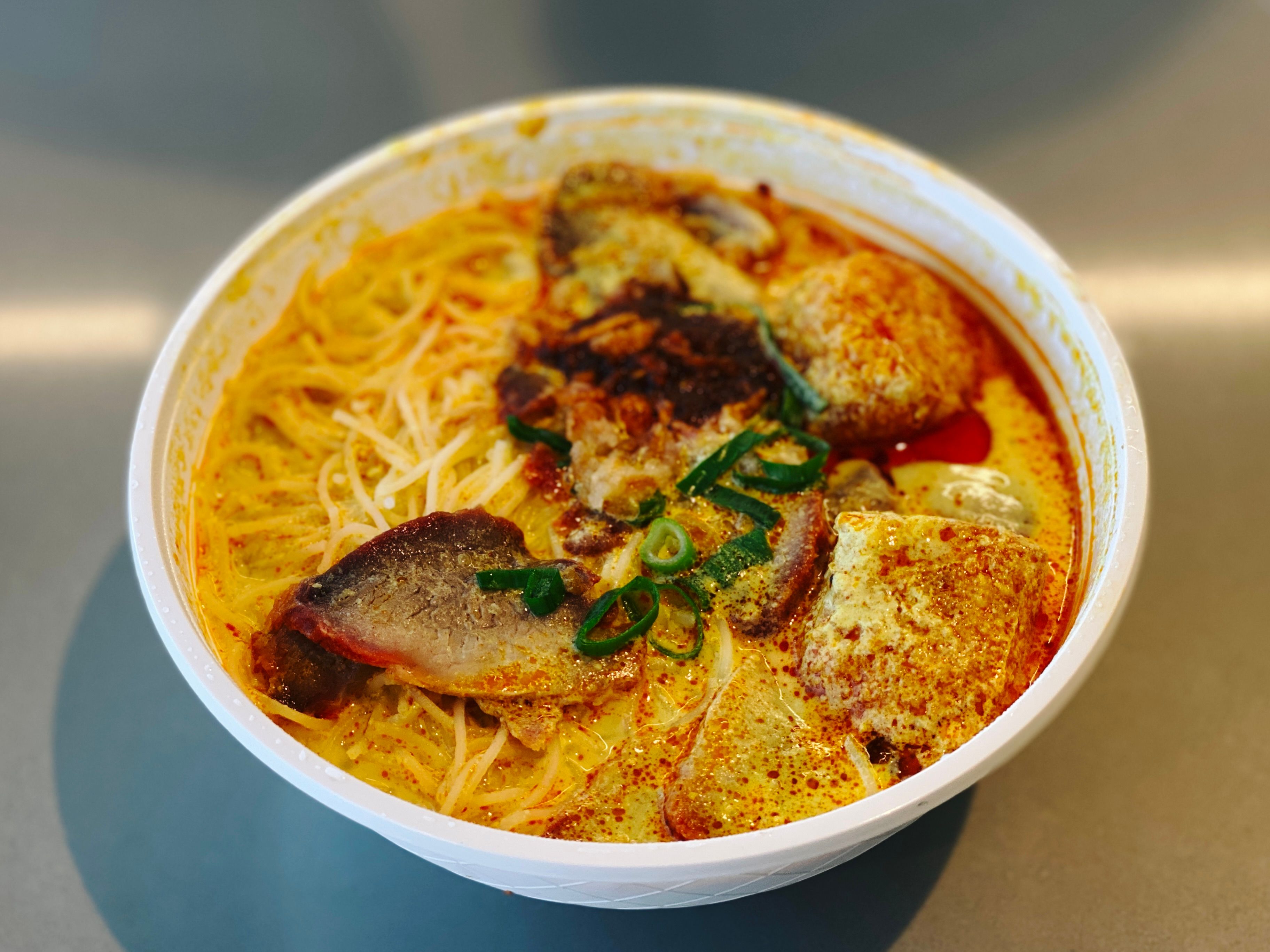 A photo of a bowl of laksa in a white takeaway food bowl. The broth is thick and rich and it's got slices of barbecue pork sitting atop the noodles.