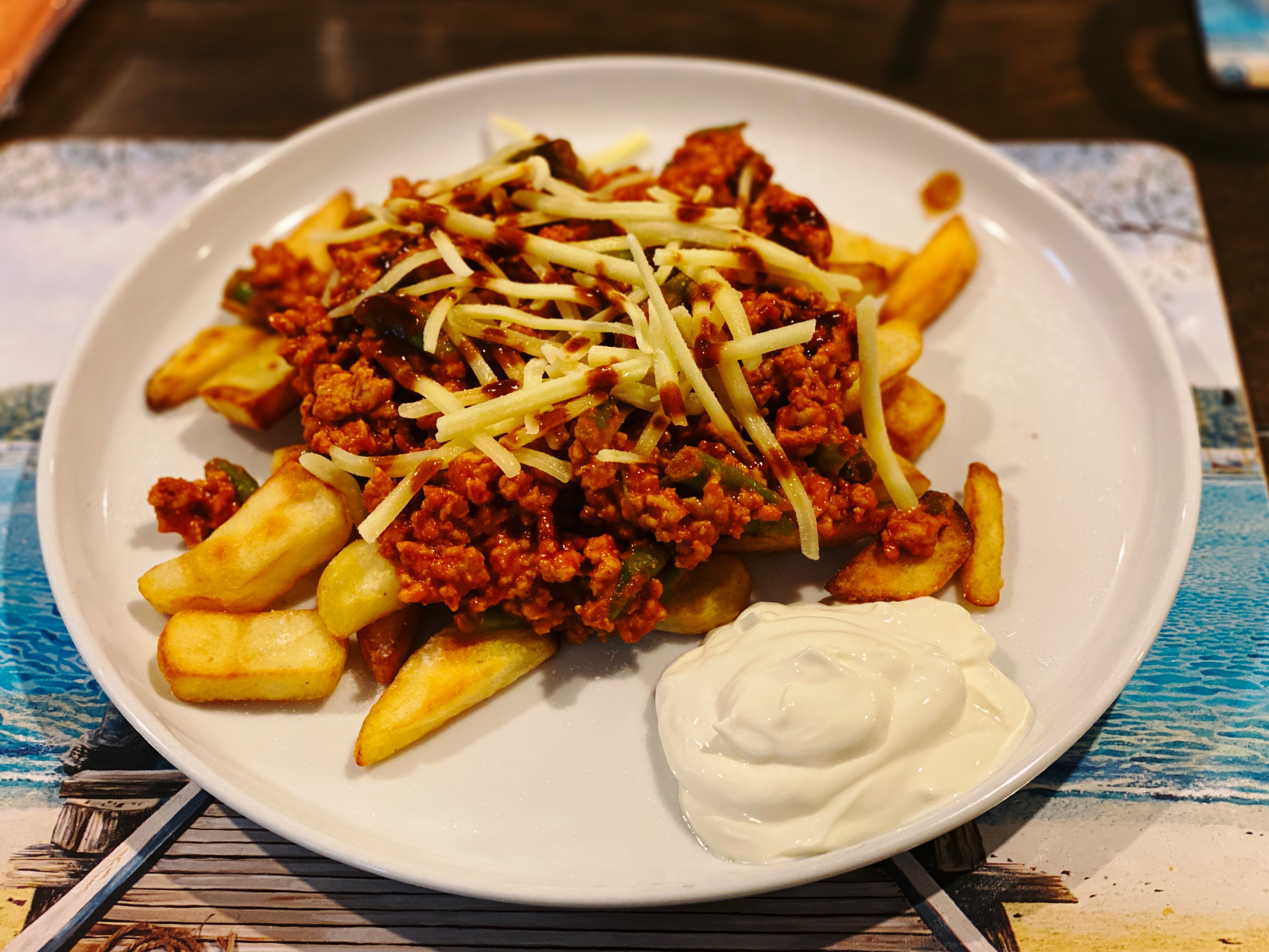 A photo of a dinner plate with a bunch of thick-cut fries covered in chilli (made with chicken mince rather than beef), with a sprinkling of shredded cheese on top and a dollop of sour cream alongside.