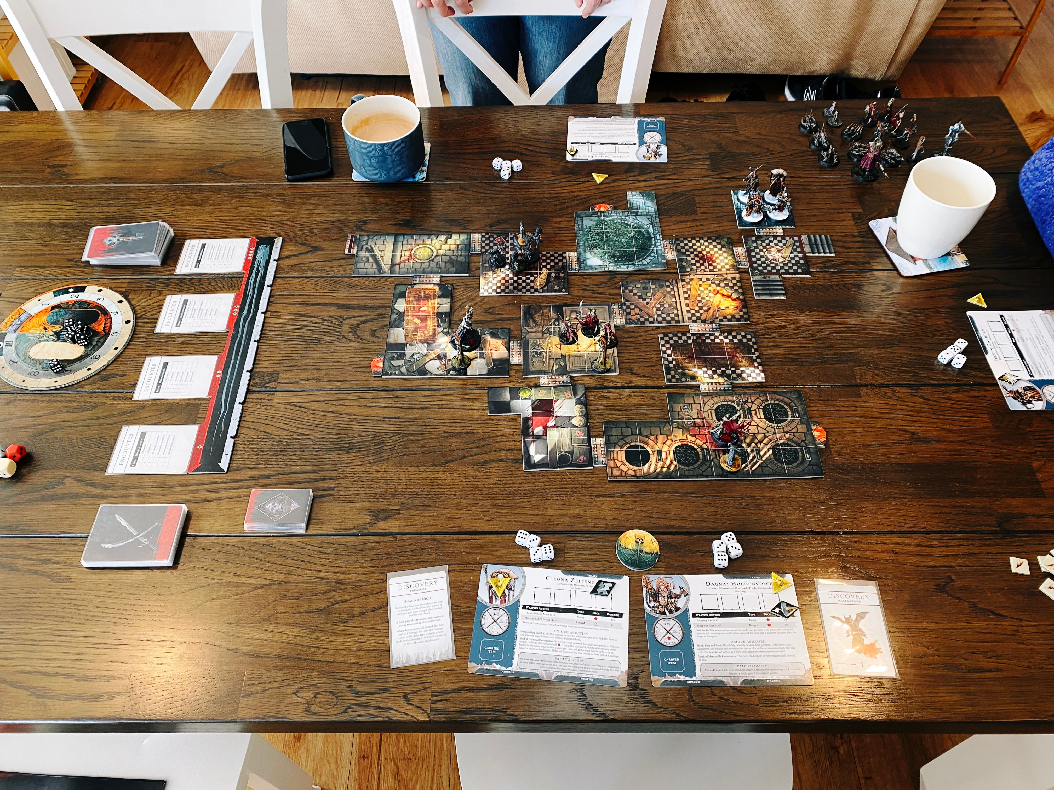 A photo of our dining table with a Warhammer Quest game set up on it. There's lots of cards and dice everywhere, and in middle are a bunch of variously-shaped tiles connected together that look like the inside of a set of buildings. There's four hero miniatures at the top (one heavily-armoured warrior woman with a sword, a vampire hunter with a long leather coat and a big hat, a steampunk dwarf, and a priest with a rich red robe and a big staff) plus four groups of hostile miniatures at various points on the board (two skeletal warriors, another larger nastier-looking skeletal warrior, three swarms of bats, and a freaky-looking extremely pale and gaunt humanoid with a big spade who's just unearthed a zombie).