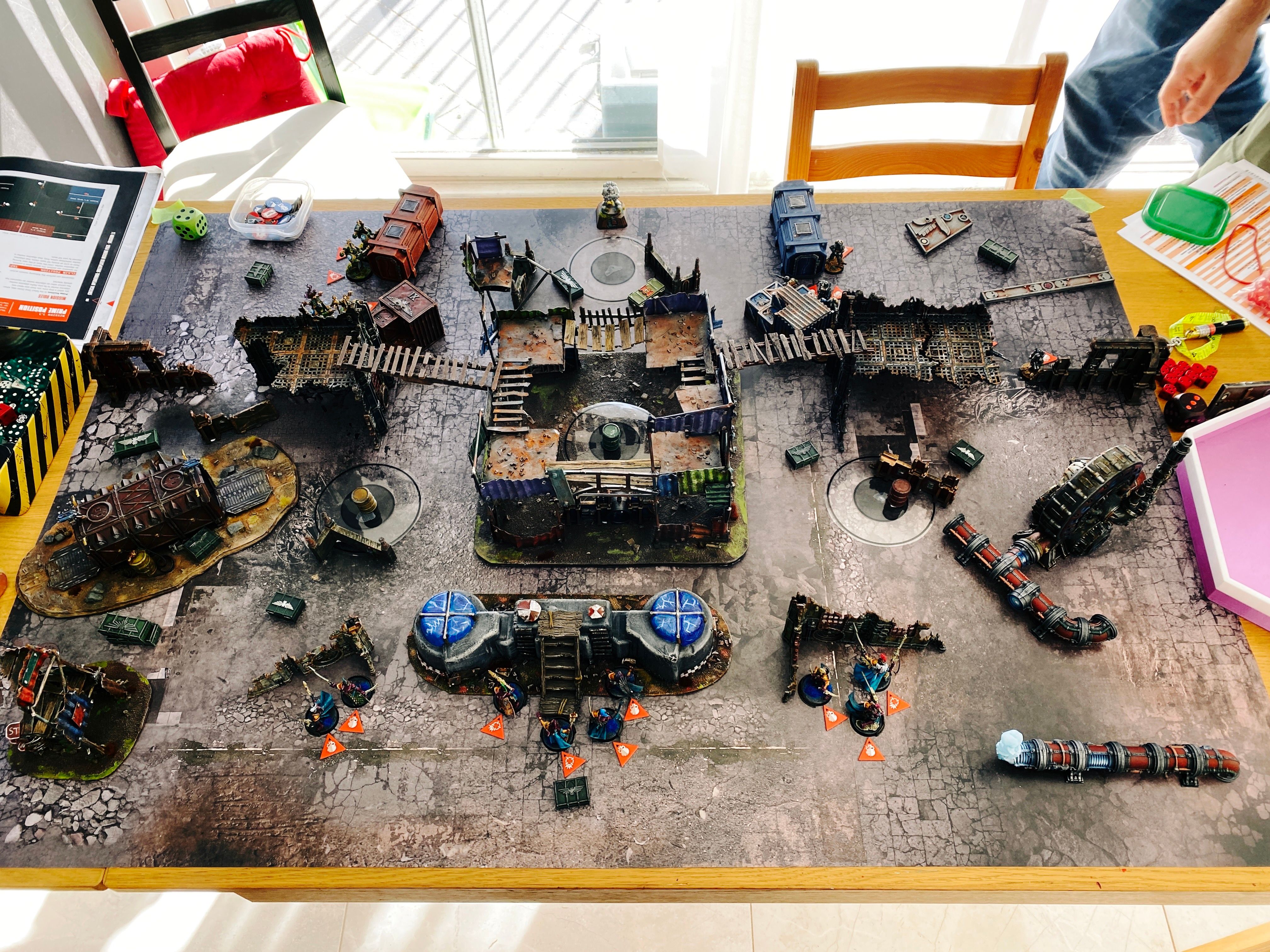 A photo of the board at the start of a game of Warhammer 40,000: Kill Team. The terrain is all rusty-looking and generally ruined, with several very ramshackle structures. Closest to the camera are my Coirsair Voidscarred, basically lithe space elves in fuchsia armour and turquoise cloaks, at the back right are the Veteran Guardsmen, a bunch of regular human warriors with big overcoats and all wearing gas masks, and at the back left are the Warp Coven, big heavily armoured super-human Chaos Space Marines whose armour is green with gold trim and they've got a very Egyptian feel to them.