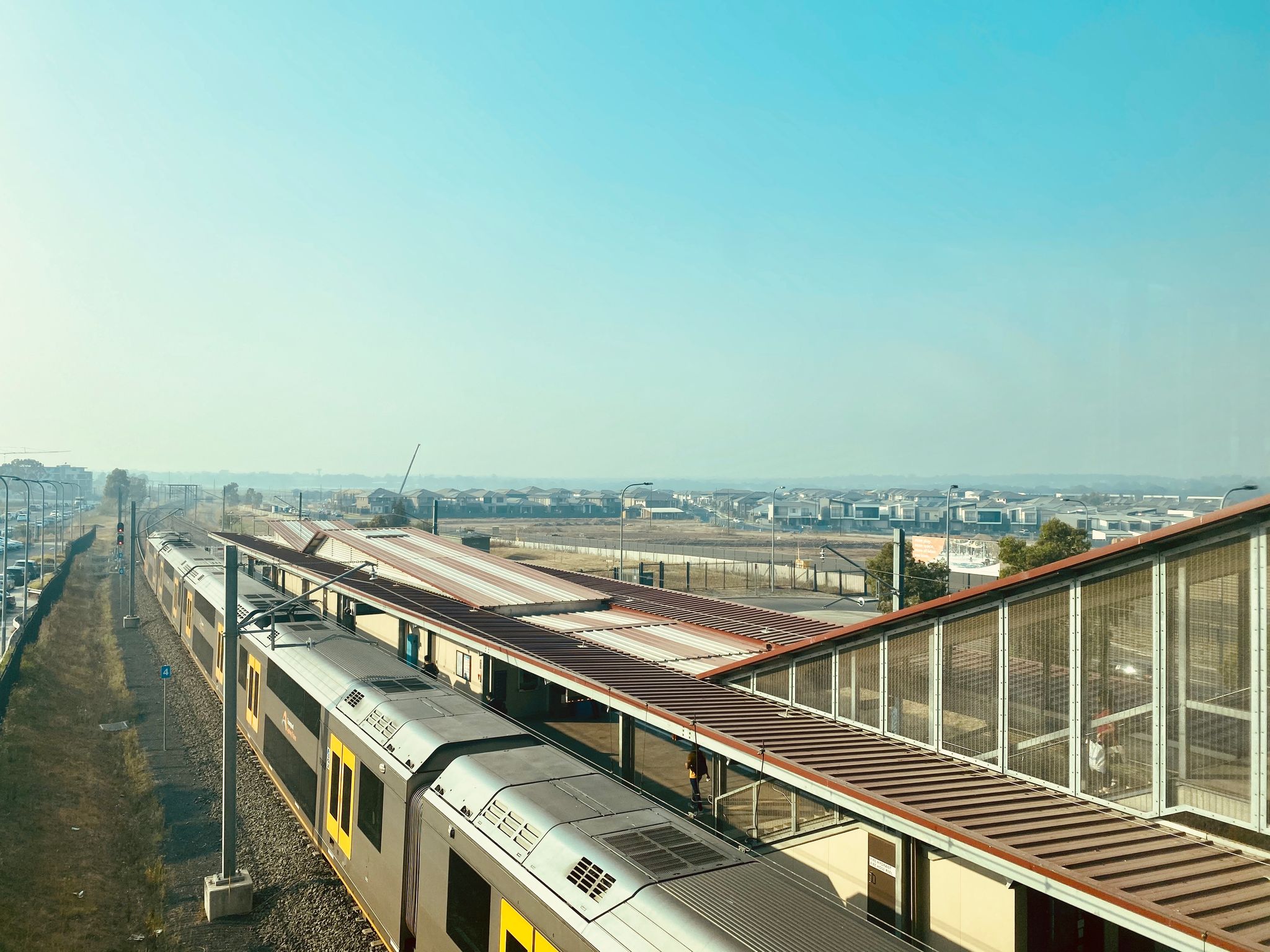 A photo taken from a train station, where the pedestrian bridge crosses above the tracks. A train is waiting at the platform below and left of frame, in the distance are houses, and the whole distance is totally hazy from bushfire smoke.