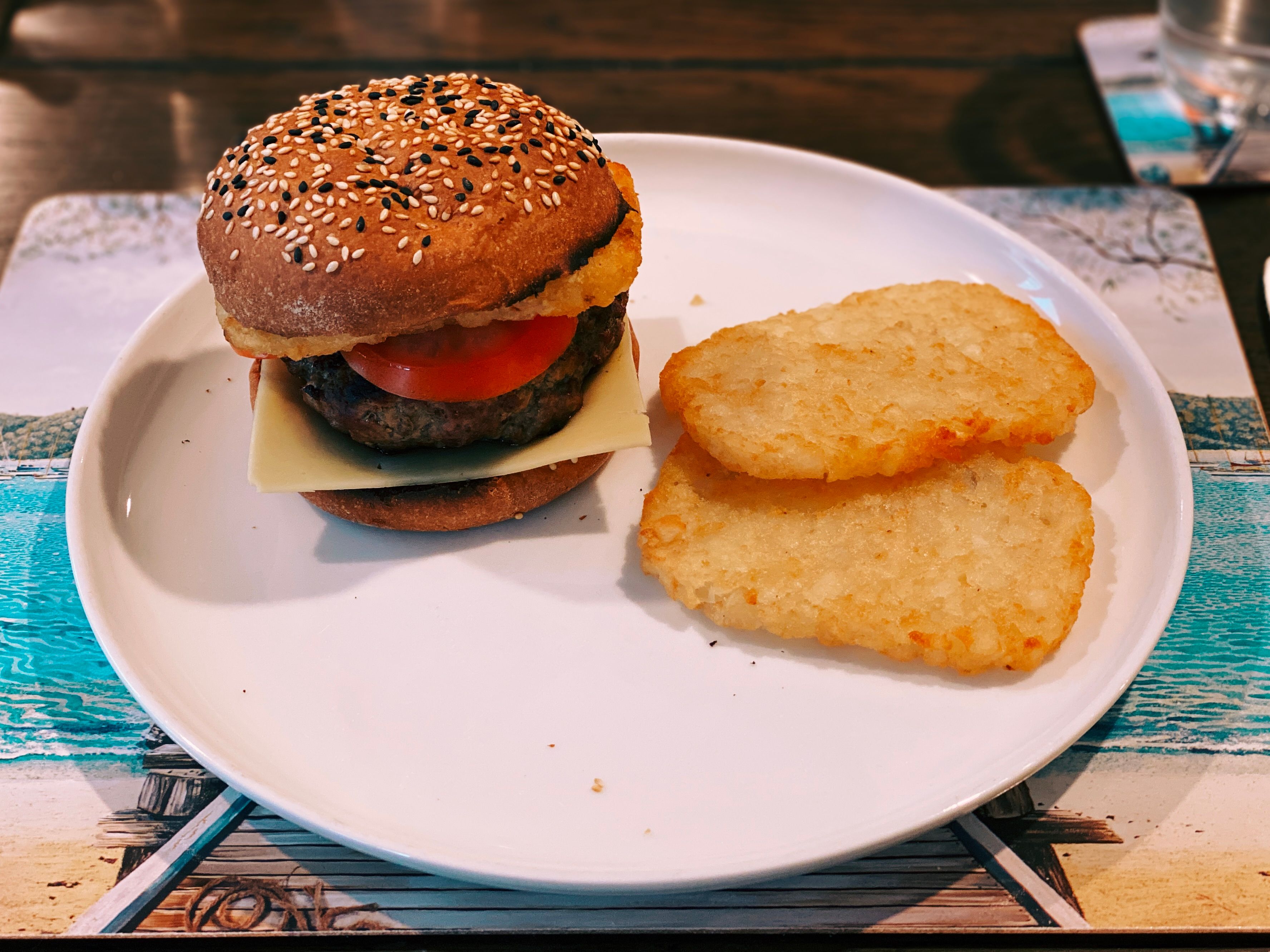 A photo of a beef burger on a plate along with two hash browns. The burger has ketchup, a slice of cheese, several slices of tomato, and one of the hash browns on it.