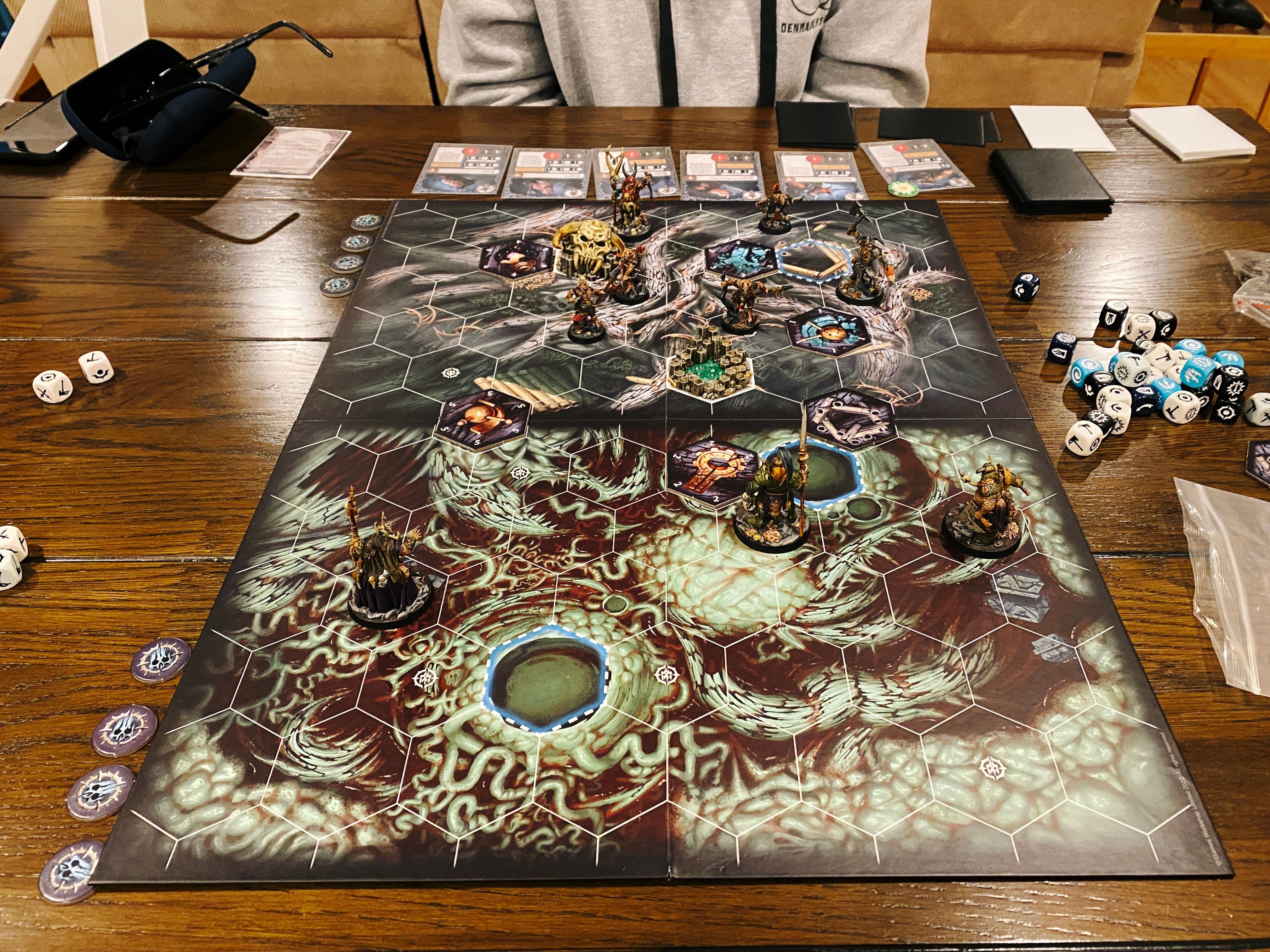 A photo of a Warhammer Underworlds game board. It has hex tiles on it, and the artwork looks like the bottom of a forest with gnarled roots everywhere. Closest to the camera are the three miniatures of The Wurmspat, they're all humanoid but corpulent with pustulant skin and extra tentacles coming from various places. At the other end of the board are the six goatmen of Grashrak's Despoilers: two are larger and more intimidating looking, one is holding a giant axe in the air and is gripping the head of someone he just decapitated, and the other four are smaller and shabbier, wielding bows and spears.