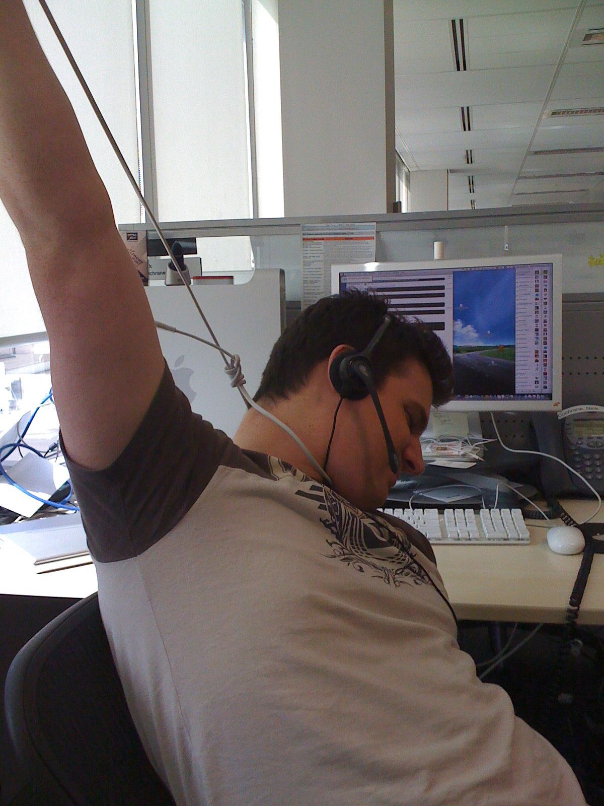 A photo of a white man sitting at his desk with a call centre headset on, his head slumped to the side and a "noose" made out of ethernet cable around his neck, holding the other end of the noose up with one hand.