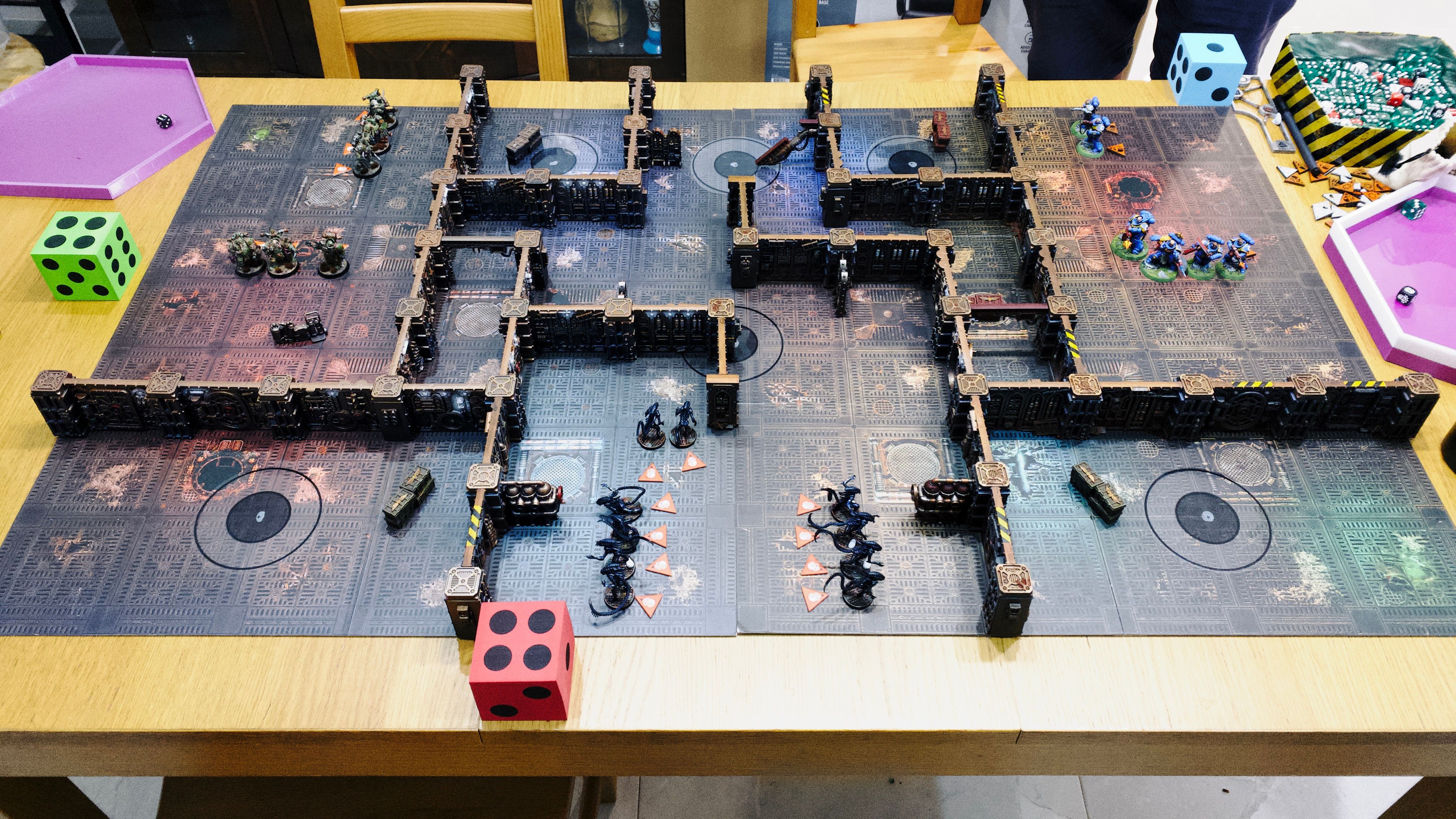 A photo of a game of Warhammer 40,000: Kill Team. The board looks like the metal of the inside of an ancient steampunk starship, and the terrain is a bunch of walls all connected together in the same steampunk style to form number of interconnecting rooms. Closest to the camera the xenomorphs from Aliens pretending to be Genestealers, painted in a very dark blue colour. On the left are the six hulking and putrid Death Guard in large military green amour with all sorts of tentacles and demon faces coming out of the armour. On the right side are six Ultramarines of broadly the same shape armour as the Death Guard, but it's a rich blue and doesn't have tentacles and demon faces in it.