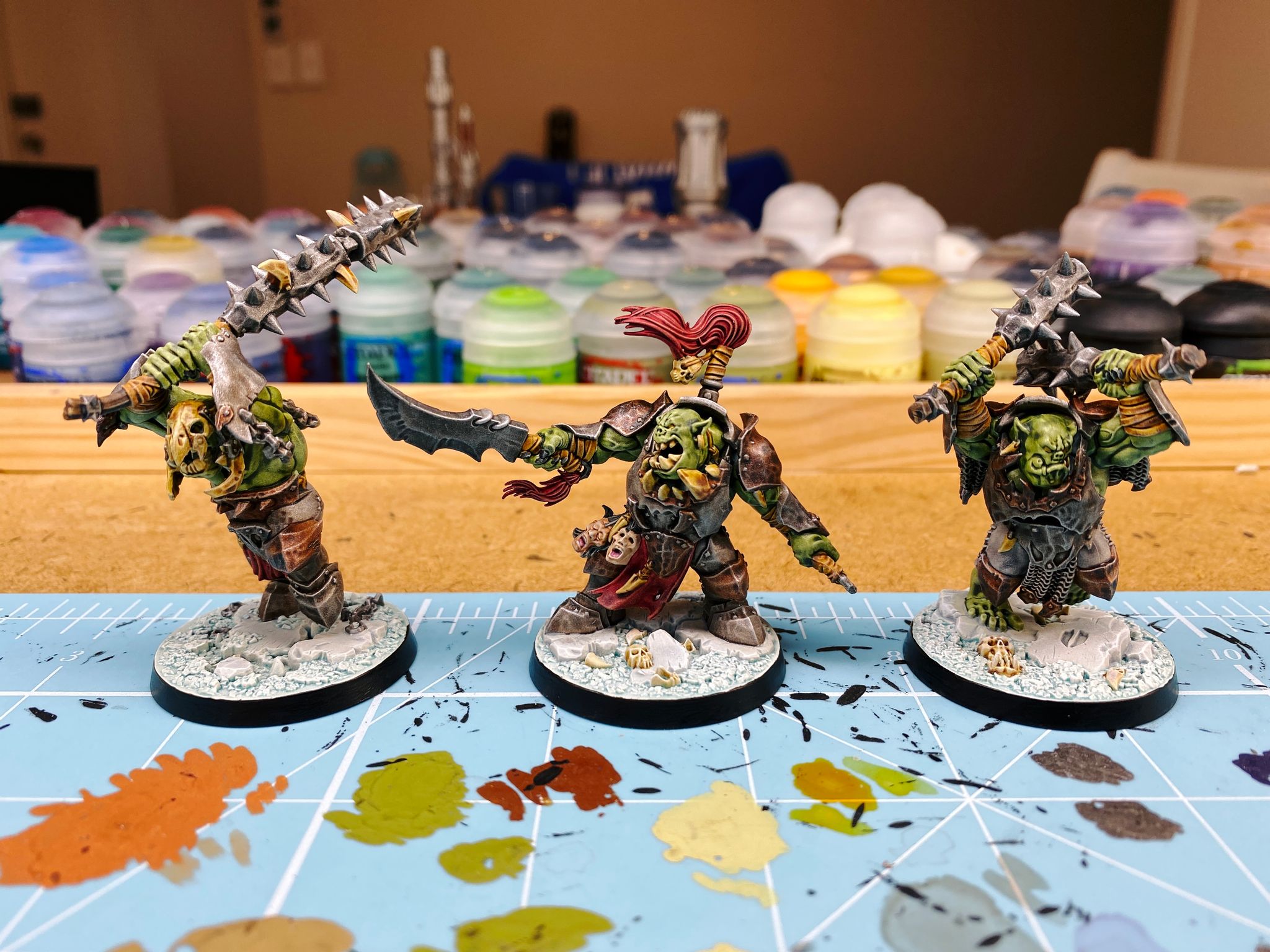 A photo of three fully painted "orruks" (Games Workshop speak for orks). They're all in battered-looking dark metal and bronze armour, the leader is pointing a sword and roaring, and the other two are mid-weapon swing. One mid-swing one is holding two metal clubs with spikes coming out of them, and the other's is the same club-looking things but joined together in a nunchuck-like fashion. Their bases are pale blue and white like snow.
